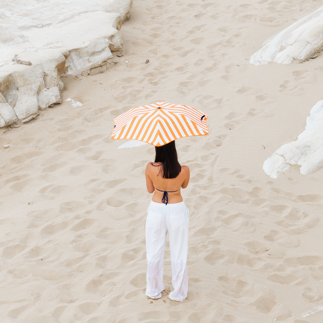 Person holding a striped umbrella on a sandy beach with rocks.