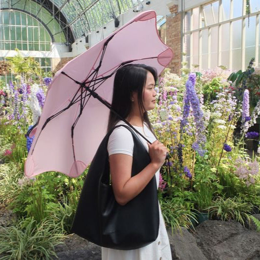 Woman holding a pink umbrella in a garden setting with flowers and greenery.