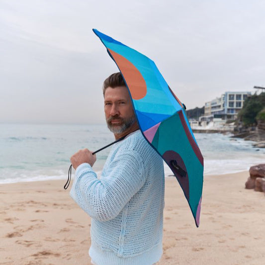 Man holding a colourful umbrella on a beach with ocean and buildings in the background